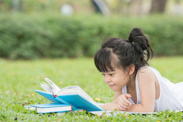 Beautiful smiling teenage girl in white blouse lying on grass and read book, against green of summer park.