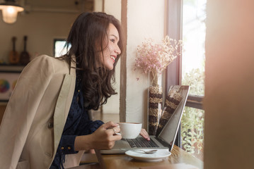 Young businesswoman sitting in coffee shop at table in front of laptop and drinking coffee. On background white brick wall and window. Girl shopping online, blogging, checking email.Online education.