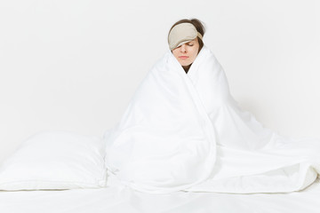 Fun tired young woman sitting in bed with sleep mask, sheet, pillow, wrapping in blanket isolated on white background. Beauty female spending time in room. Rest, relax, good mood concept. Copy space.