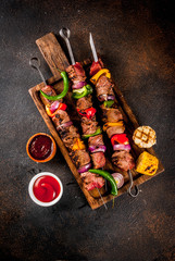 Wall Mural - Fresh, home-cooked on the grill fire meat beef shish kebab with vegetables and spices, with barbecue sauce and ketchup, on a dark background on a wooden cutting board above copy space