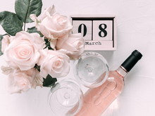 8 March. Happy Women's Day. Wooden Block Calendar. White Rose, Flowers. Bottle Of Pink Wine And Two Glasses Of Wine On A Soft Light Pink Wood Table, Flat Lay, Top View, Copy Space. Selective Focus 
