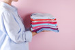 © Evrymmnt - Close up of young woman wearing blue cotton shirt holding stack of perfectly folded multicolor shirts. Female w/ pile of different color clothing in her hands on pale pink background. Laundry concept.