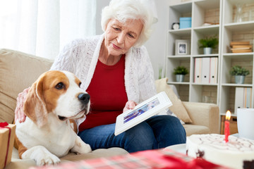 Wall Mural - Portrait of white-haired senior woman holding photograph and remembering husband while sitting in comfortable armchair at home with her dog, copy space photo in frame by me 