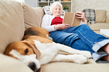 Wall Mural - Full length portrait of white haired senior woman resting on couch reading book with sleeping dog enjoying afternoon nap at home, copy space