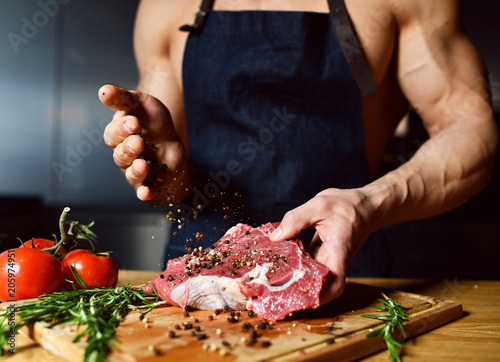 Fotografie, Obraz Strong sport man hands pour pepper prepare cook beef steak ribs on dark kitchen