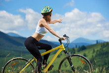 Attractive Woman Female Riding On Yellow Mountain Bicycle, Wearing Helmet, Pointing At Something In The Distance, Enjoying Valley View On Summer Day. Mountains And Blue Sky On The Blurred Background