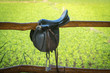 Horse saddle hanging on a wooden fence with green background.