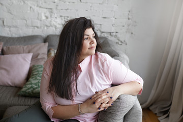 Wall Mural - Indoor shot of beautiful plus size fashion female model with straight black hair relaxing on comfortable gray sofa by the window at home, having pensive thoughtful expression, wearing pink shirt.