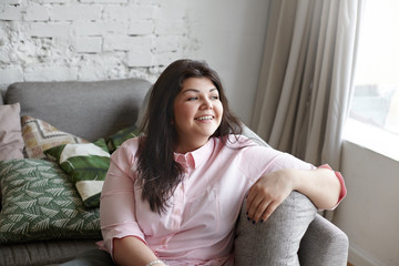 Wall Mural - People, lifestyle, bodypositivity and obesity concept. Portrait of cheerful charming young overweight woman with beautiful features smiling, posing indoors, sitting on sofa, looking though window