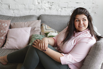 Wall Mural - Horizontal portrait of charming body positive plus size adorable young woman sitting on sofa with her legs bent, dressed in black jeans and pink shirt, feeling carefree while relaxing at home