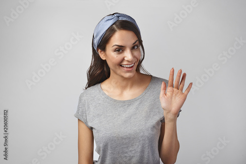 Horizontal studio shot of attractive friendly looking young brunette female with headband smiling happily, saying Hello, Hi or Bye, waving hand at camera. Greeting, goodbye and body language