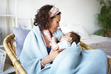 Wall Mural - Childcare, parenting, nursing and lactation concept. Portrait of beautiful young mixed race woman with curly hair sitting in chair covered with blue blanket and breastfeeding her cute sweet baby son