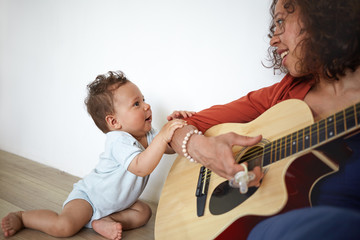 Happy young Hispanic female musician sitting on floor, playing guitar to her adorable cute baby son who is singing along with her, Music, art, talent, creativity, infancy, education and childcare