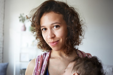 Indoor shot of beautiful young brown eyed mixed race housewife posing at home, carrying her nine month old son in baby sling. Motherhood, childcare, parenting, nursing and maternity concept