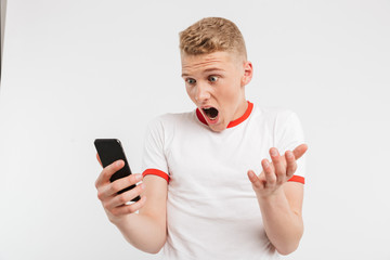 Poster - Photo of outraged young man wearing casual style screaming while holding and using smartphone, isolated over white background