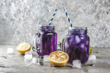 Healthy summer cold beverage, iced organic blue and violet butterfly pea flower tea with limes and lemons, grey concrete background copy space top view