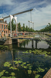 Fototapeta Pomosty - Bascule bridge over the canal with aquatic plants and brick houses on a sunny day in Weesp. Quiet and pleasant village full of canals and green near Amsterdam. Northern Netherlands.