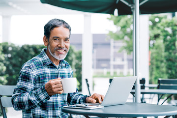 Portrait of happy white stylish short beard mature man drinking coffee while using laptop. Casual urban lifestyle of retired hispanic people, adult asian man sitting, smiling at modern cafe outdoor.