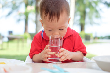 Wall Mural - Cute little Asian 18 months / 1 year old toddler boy child sitting in high chair holding & drinking glass of water by himself at restaurant in beach resort, best beverages for child's health concept