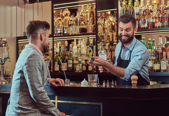 Stylish brutal barman in a shirt and apron makes a cocktail at bar counter background.