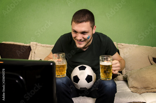 Fotografie, Obraz Young adult guy sitting with two beer mugs and soccer ball, watching match on TV