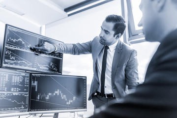 Wall Mural - Businessmen trading stocks online. Stock brokers looking at graphs, indexes and numbers on multiple computer screens. Colleagues in discussion in traders office. Blue toned black and white.