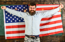 Man With Beard And Mustache On Happy Face Holds Flag Of USA, Wooden Background. American Teacher In Eyeglasses Holds American Flag. Student Exchange Program. American Educational System Concept.