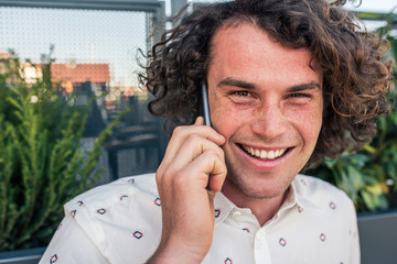 Outdoor closeup portrait of smiling bussines male with freckles in stylish shirt, enjoying call conversation on cell phone while sitting at city terrace. People, lifestyle, modern technology concept