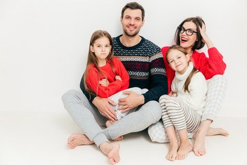 Portrait of happy family of four members: attractive brunette woman, her husband and two small daughters sit on floor, isolated on white background. Parenthood concept. Lovely family indoor.