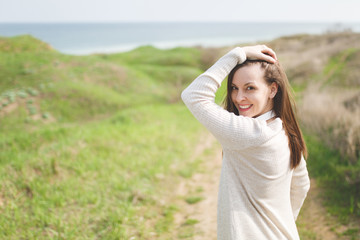 Young brunette smiling beautiful woman in light casual clothes keeping hand near head walking in sunny weather in field near water on green background. Beautiful landscape. Lifestyle, leisure concept.