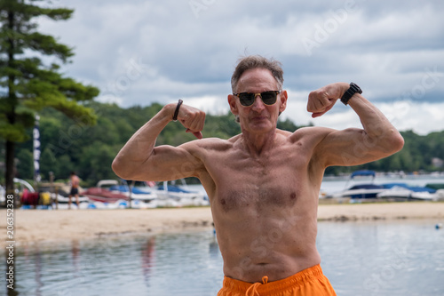 Fotografie, Obraz Old baby boomer man senior citizen in yellow bathing suit flexing his muscles with a smooth lake and beautiful beach in background