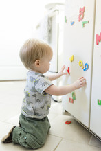 Side View Of Baby Boy Playing With Colorful Magnetic Letters And Numbers On Metallic Cabinet At Home