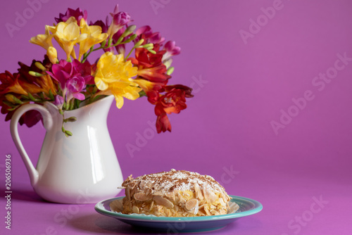 Fotografie, Obraz Portuguese pastry on a turquoise plate with a pitcher of freesia flowers behind it on a purple background