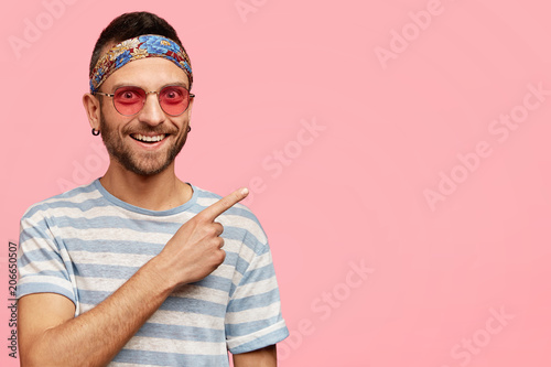Fotomural Horizontal shot of pleasant looking young hippy male with stubble, wears stylish pink sunglasses and headband, indicates at upper right corner, poses in studio