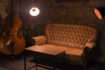 Modern jazz bar interior design, leather sofa, cello music instrument, lamps. Stone wall on background, brutal atmosphere