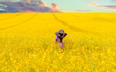 pretty young woman in violet dress and black hat on blooming sunset rapeseed field