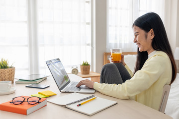 Poster - Asian female sit with knees up use social media with laptop on table and drink orange juice in bedroom at home.Work at home concept.work from home.relax lifestyle.