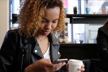 A Young Beautiful African-American Woman In A Leather Jacket With A White Paper Glass In One Hand Looks Into The Phone. Close-up Portrait. Cafe With A Window. Life Style.