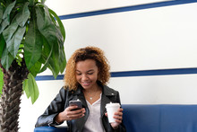 A Young Beautiful African American Woman In A Leather Jacket With A White Paper Glass Sitting On A Blue Sofa Near A Palm Tree And Looking At The Phone. Cafe Or Waiting Room At The Airport. Copy Space.