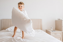 adorable toddler boy in white bodysuit playing with pillow on bed at home