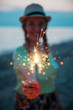 © ManuPadilla - beautiful young woman wearing braids and straw hat at the sunset beach. she is blurred with focus in the sparkler. Celebration time and happiness concept