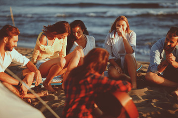 Wall Mural - Happy friends sitting on the beach singing and playing guitar during the sunset