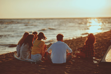 Happy friends sitting on the beach singing and playing guitar during the sunset