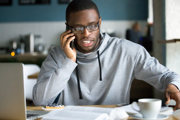 African businessman talking on phone sitting at cafe table, busy entrepreneur working distantly in coffee house with laptop papers speaking on mobile, black man making call having lunch in cafeteria