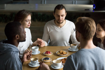 Grateful multiracial young friends holding hands sitting at home or cafeteria table saying grace concept, religious people blessing food praying meditating thanking before eating breakfast together