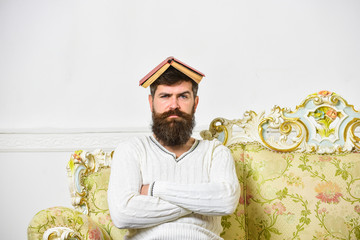 Macho sits with open book on head, like roof. Guy, teacher overdid with teaching, became crazy professor. Man with beard and mustache sits on sofa, white wall background. Overwork concept.