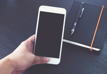 Poster - Hand holding Blank screen of mobile phone over black notebook on black wooden office desk, Mock up for display of design or content,Clipping path smart phone screen.