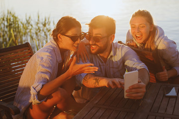 Two young woman and man using smartphone by the river and smiling