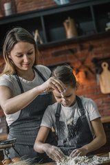 Canvas Print - Cute girl and mother in aprons using flour to cook the dough on kitchen at home. 