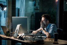 Man Covering Mouth While Yawning At Table In Dark Office Having Overtime. 